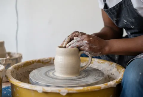 Close-up of hands molding a ceramic cup on a pottery wheel with clay-covered fingers against a white background