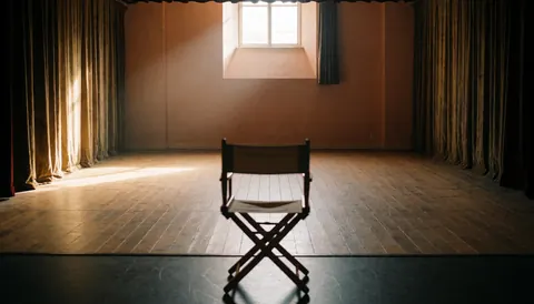 An empty theater stage featuring a director chair in the center, natural light from a window, velvet curtains, and a wooden floor.
