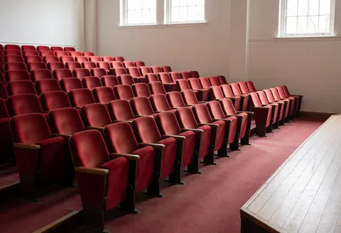 View of an empty theater auditorium featuring rows of red seats facing a wooden stage, illuminated by natural light from high windows.