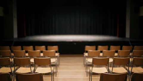 An empty theater stage featuring rows of wooden chairs and a dark curtain backdrop under dim lighting, suitable for professional text overlays.