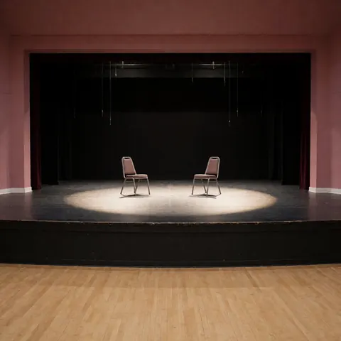 Two empty theater chairs placed under a central spotlight on a stage with pink walls and black curtains in the background