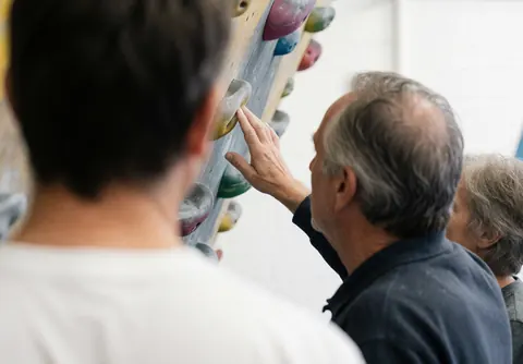 Climbing instructor points to a hold on the wall while a student watches from the foreground in a bright gym.