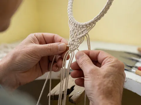 Close-up of adult hands working with white cord on a pale yellow background, showing the texture of the macrame knots and braids in progress