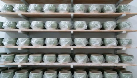Wooden shelves holding green ceramic pots wrapped in clear plastic against a soft, diffused background with cool, even lighting and terracotta tones