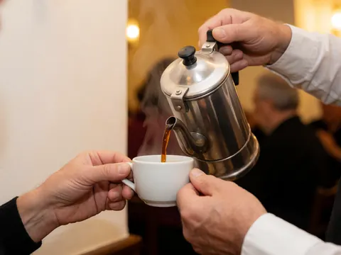 Close-up of a hand pouring hot coffee from a metal percolator into a white ceramic cup against a warm tango-themed background.