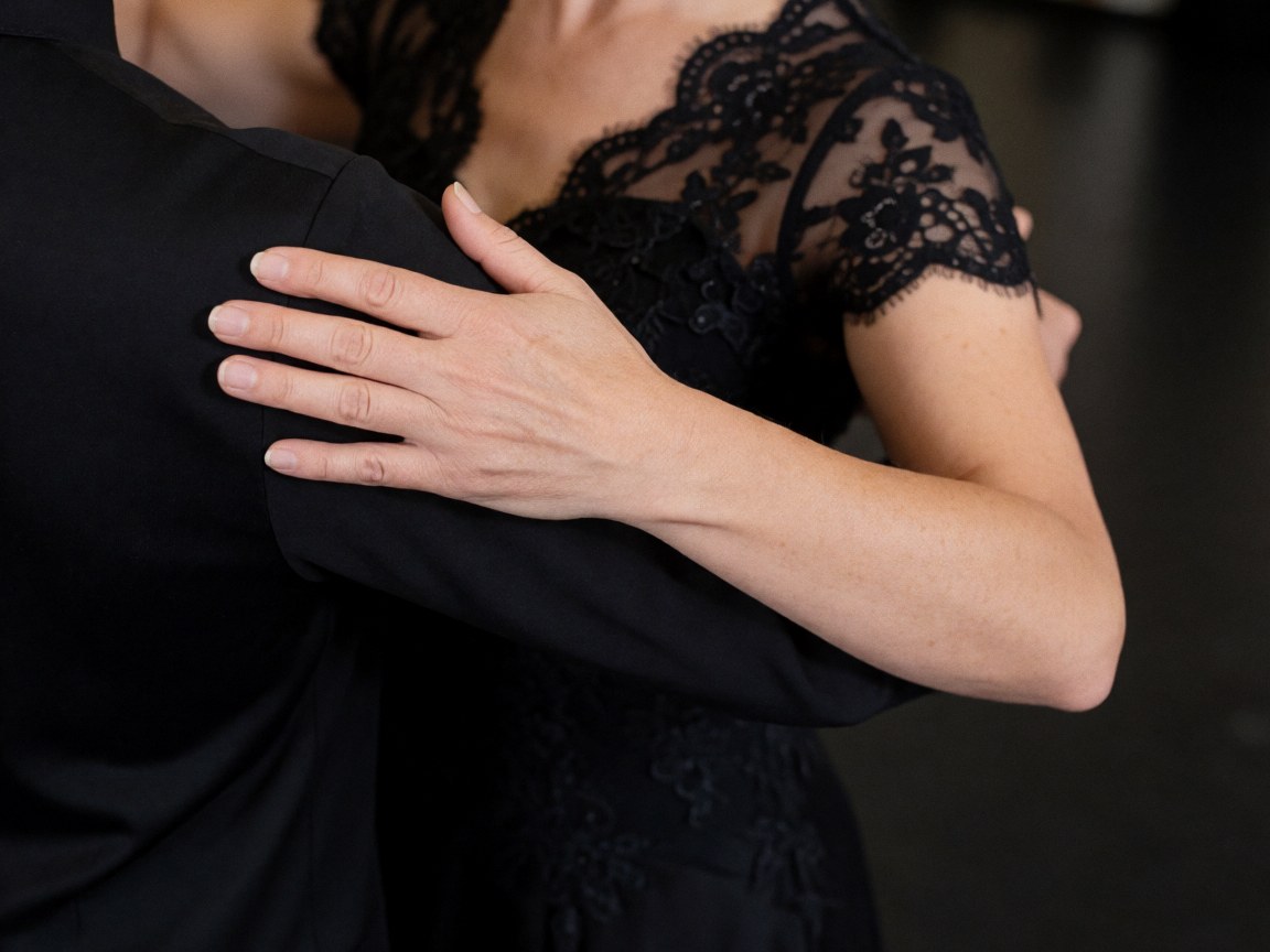 Close-up of a woman's hand in black lace fabric gripping her partner's shoulder during a tango dance pose against a dark blurred background.