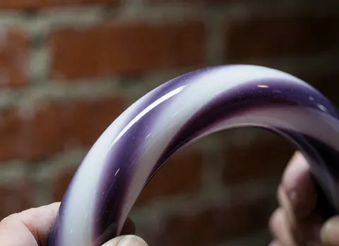 Close-up of a polished purple and white gradient glass ring held against a red brick background, highlighting artisanal craftsmanship and glossy reflections.