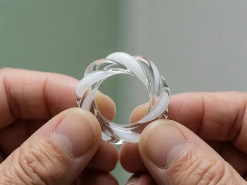 Close-up of a braided glass ring held in hands against a soft gray background with a pale green wall, highlighting artisanal craftsmanship details