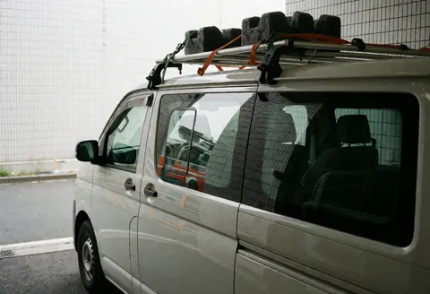 White service van equipped with a roof rack for transporting glass, parked near a metal mesh wall with reflections visible in the windows.