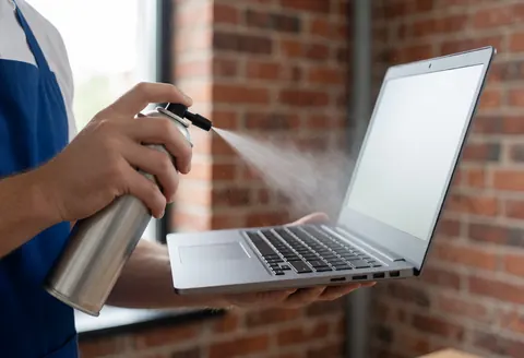 Technician in blue apron cleaning laptop ventilation with compressed air, dust particles floating, red brick wall background