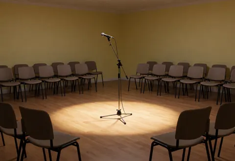 An empty recital stage featuring a central microphone, audience chairs, and warm lighting on the wooden floor.