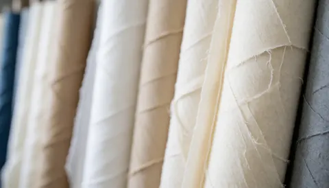 Stacks of neutral colored fabric rolls stored in a warehouse, providing a soft textured background suitable for upholstery business materials.