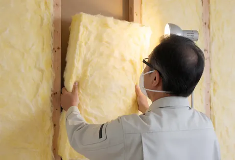 A worker wearing a mask and safety glasses installs a yellow thermal insulation panel between wooden beams during construction.