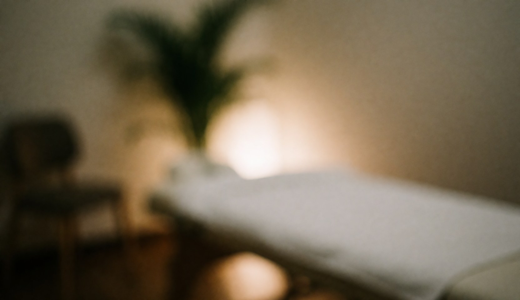 A covered massage bed with a blurred plant in the foreground against a warm, earthy-toned background suitable for text overlay.