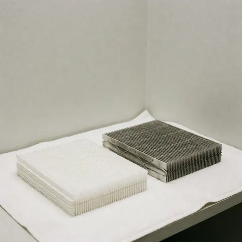 A clean white heating filter and a dirty used one placed on white fabric against a light gray wall corner to show the need for replacement.