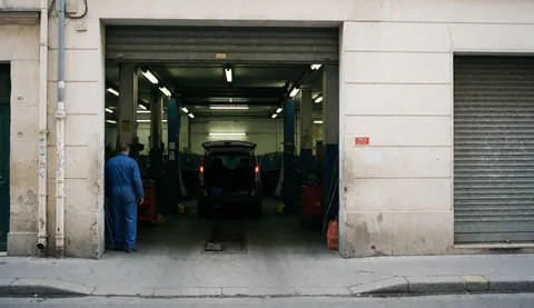 Mechanic in blue workwear standing near the open garage entrance with a vehicle inside and workshop lights on
