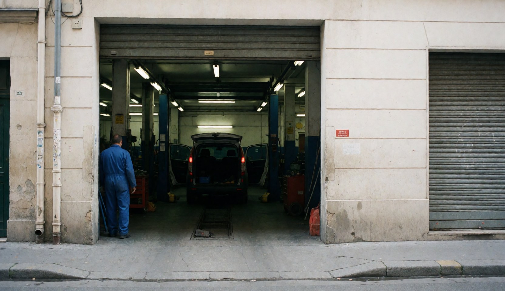 Mechanic in blue workwear standing near the open garage entrance with a vehicle inside and workshop lights on