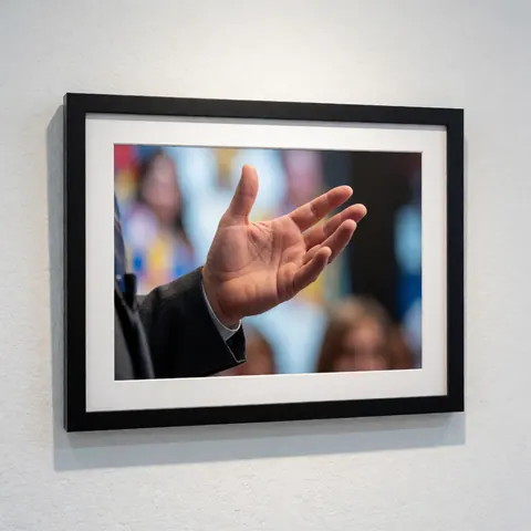 A framed photograph of a hand making a gesture, displayed on a white textured wall with a blurred background of people.
