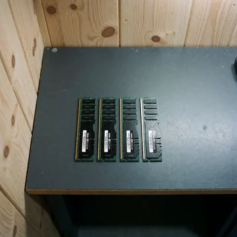 Four DDR4 RAM memory modules with white labels arranged in a row on a dark work table with light wood walls in the background