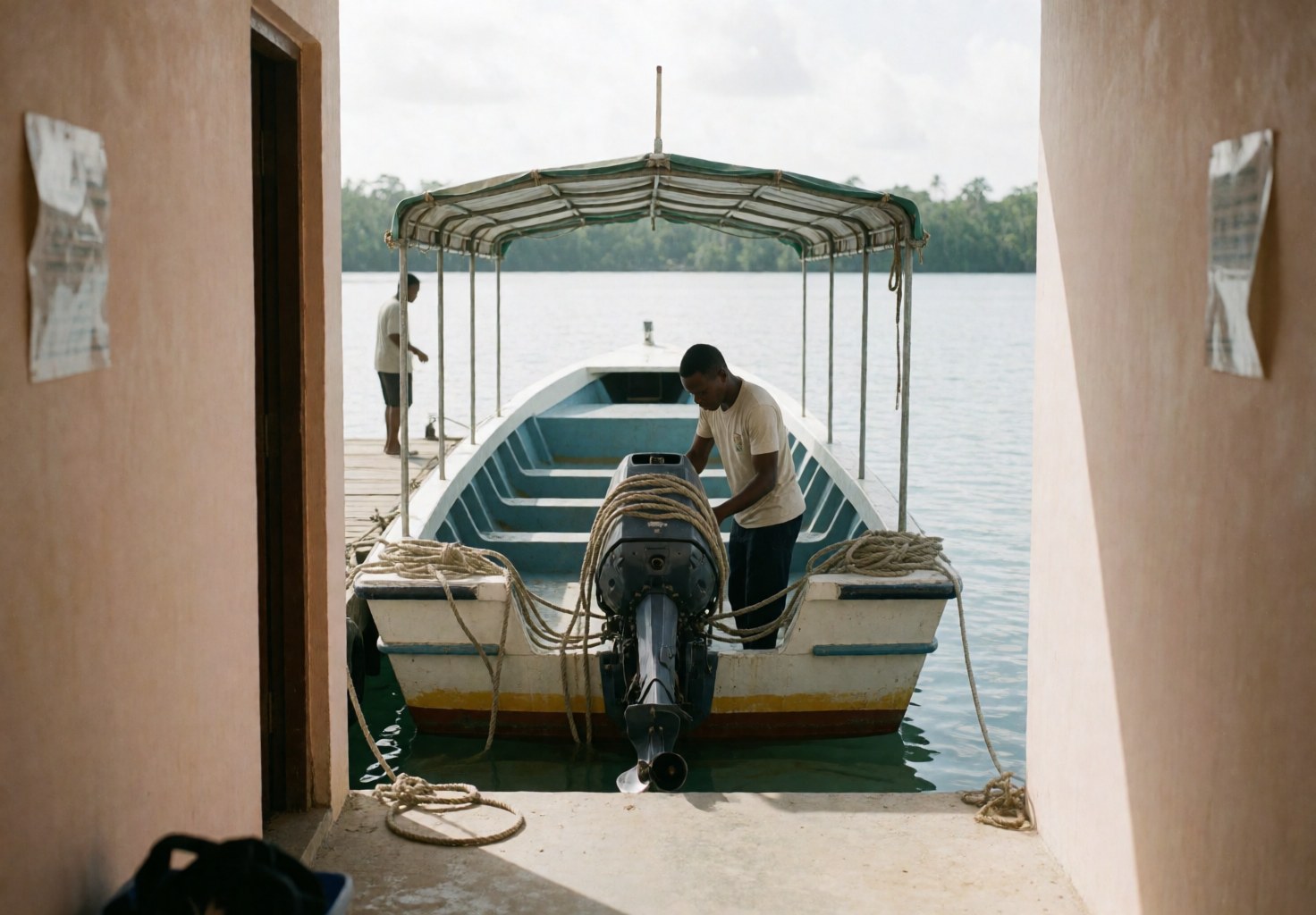 A worker adjusts the engine of a tour boat moored at a dock with a canopy and ropes, reflecting calm water and tropical vegetation.