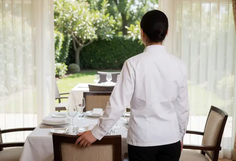 Waitress with back to camera in clean restaurant with set table, white curtains, and garden view
