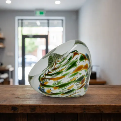 Handcrafted glass weight with green and yellow patterns resting on a rustic wooden counter in a glassblowing workshop