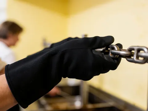 Close-up of a hand wearing a black glove tightening a thick metal chain with a blurred background of light yellow walls.