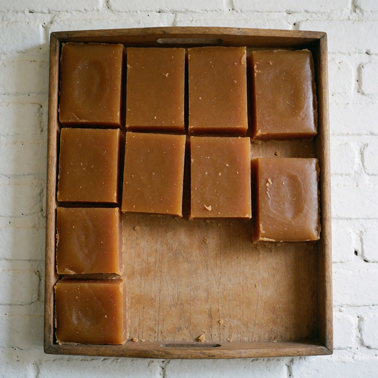 Artisanal panela pieces on a wooden tray against a white brick wall with natural lighting and visible crystallization texture