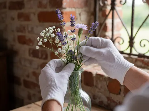 Hands wearing white gloves place wildflowers into a clear glass vase against a red brick wall and wrought iron frame.