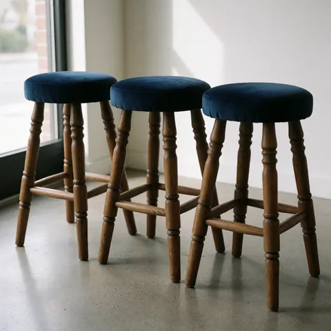 Three restored stools featuring dark blue velvet seats and polished wooden legs, set against a natural concrete background with soft window light.