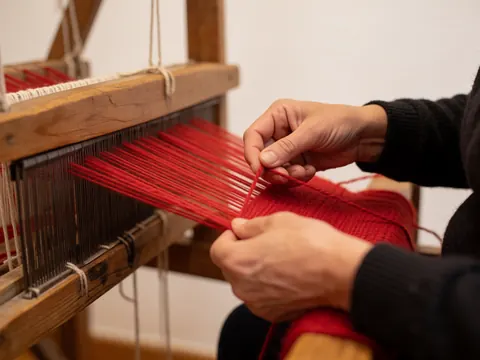 Close-up of hands weaving red wool on a rustic wooden loom with warm lighting against a white background, suitable for rural lodging details.