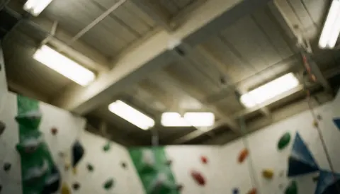 Out of focus ceiling of a climbing gym featuring green and red handholds, exposed beams, and fluorescent lights, suitable as a text background.