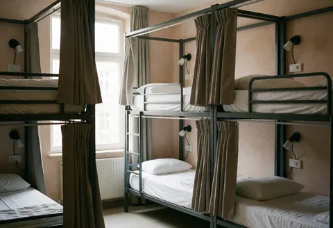 Hostel dormitory featuring bunk beds with curtains, wall lights, and outlets against terracotta walls, illuminated by natural light from a window.
