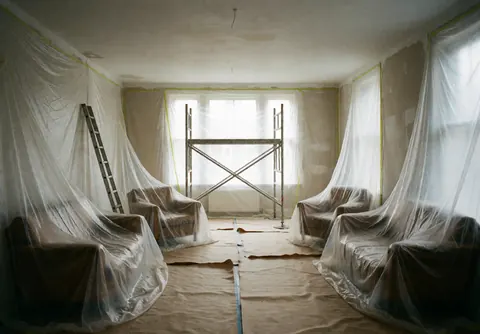 Empty room with furniture covered in plastic, scaffolding in the center, and kraft paper on the floor, lit by natural light from windows.