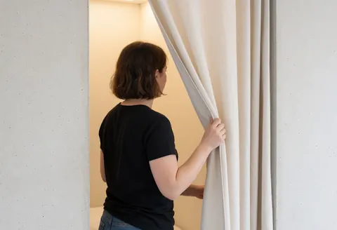 Woman closing white curtain in private hostel room with concrete walls and warm interior light