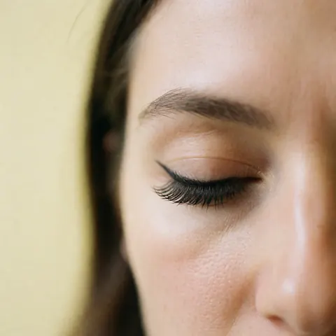 Close-up of an eye featuring natural black eyelash extensions against a pale yellow wall with soft lighting and visible texture.