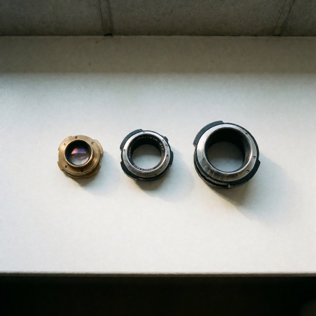 Three vintage lens adapters, one bronze and two black metal, arranged in a row on a white surface with natural light and soft shadows