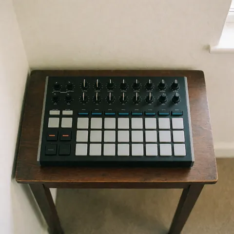 MIDI controller with pads and knobs on a dark wooden table against white walls with soft natural lighting