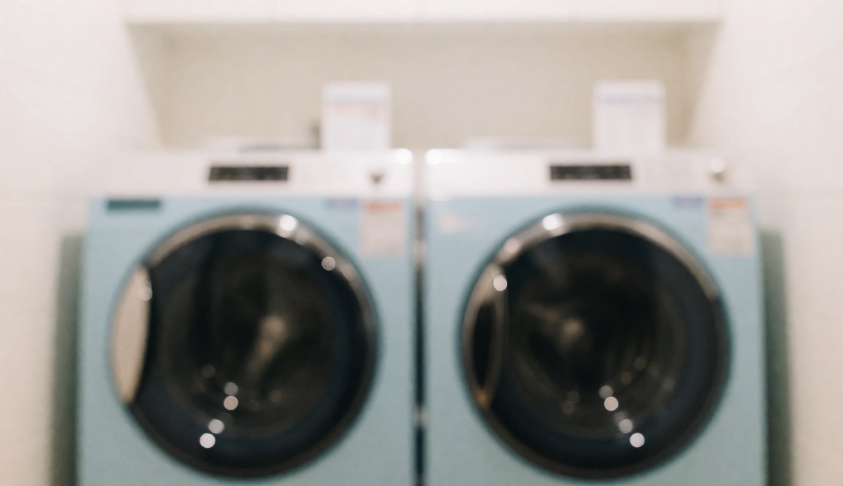 Out of focus background featuring two light blue washing machines in a laundry setting, suitable for text overlays regarding cleaning and laundry services.