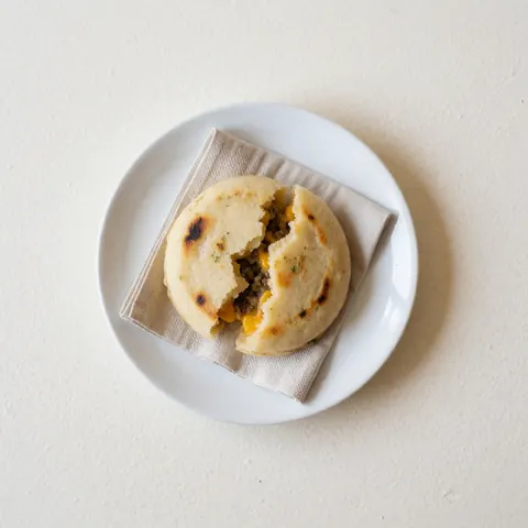 Hot arepa with melted cheese and meat served on a white ceramic plate atop a linen cloth with bright, clean lighting.