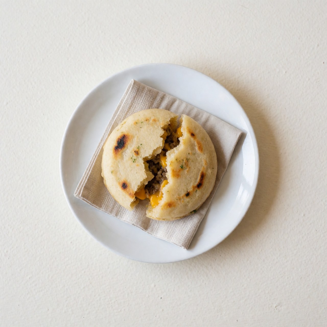 Hot arepa with melted cheese and meat served on a white ceramic plate atop a linen cloth with bright, clean lighting.