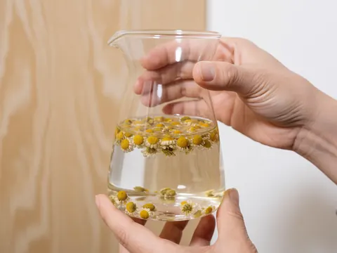 Hands holding a glass jar filled with hot chamomile tea, showing bubbles and petals against a wooden background and white wall