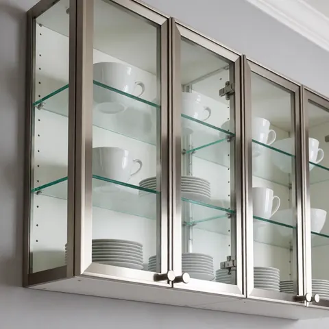 Modern kitchen cabinet featuring glass doors and stainless steel frames, displaying white dishes and cups on glass shelves.