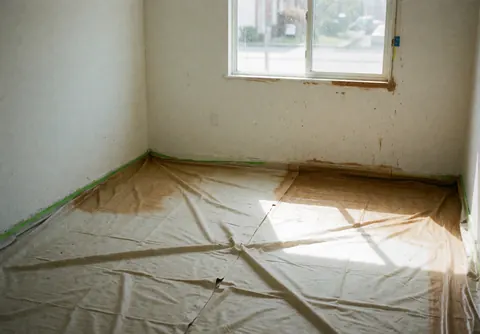 Room with white walls and floor protected by plastic for painting, natural light from window, clean space ready for work.