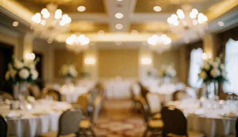 Decorated event hall background featuring round tables with white tablecloths, chairs, and blurred chandelier lights, ideal for text overlay and design projects.