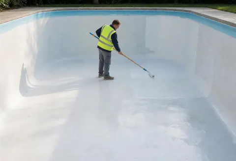 Technician in safety vest using a long roller to apply white waterproofing coating on the interior wall of an empty swimming pool.