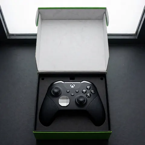 Black Xbox Elite controller with silver accents inside its retail packaging on a dark gray surface with natural overhead lighting