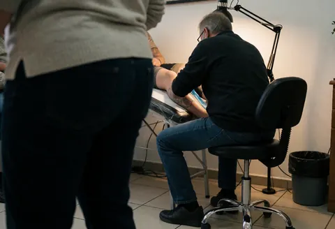 Tattoo artist wearing glasses and a black shirt sits in a chair, working on a client's leg under a desk lamp in a tiled studio.