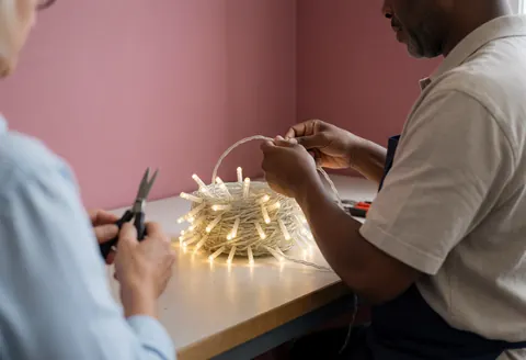 Two individuals work in a workshop with a pink wall, one rolling up camping lights while the other holds scissors, showing organized preparation.