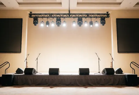 Empty event stage with lights, microphones, speakers, and large screens, beige walls, ready for presentations.